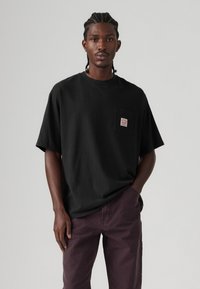 Black cotton T-shirt with a crew neck and short sleeves, featuring a chest pocket with a label. Paired with dark purple pants.