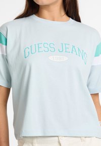 Šviesiai mėlyna medvilninė marškinėlių su apvalia iškirpte, ant kurios yra užrašas "GUESS JEANS 1981" turkio spalvos raidėmis, o rankovės su baltomis ir turkio akcentais yra spalvoti.