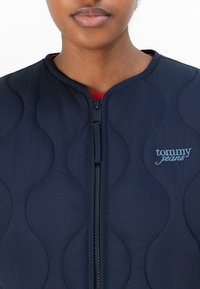Marinblå vadderad jacka med rund halsringning, med en hel dragkedja och "tommy jeans" broderat i ljusblått. Strukturerad yta med scallop-mönster.