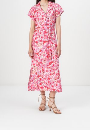 Kobieta ubrana w różowo-czerwoną kwiecistą sukienkę midi z krótkimi rękawami, kołnierzykiem, wiązaniem w pasie oraz beżowe sandały na obcasie z paskami.