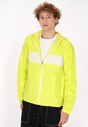 Jachetă ușoară, galben-neon, cu o dungi orizontale albe, cu fermoar în față, glugă și manșete elastice. Modelul o poartă peste un tricou alb.