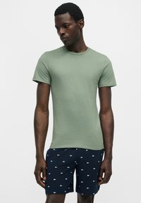 Homme portant un t-shirt vert clair à manches courtes et un short bleu marine avec des logos crocodiles blancs sur un fond blanc uni.