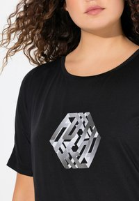 Černé tričko s velkým geometrickým stříbrným grafickým designem na hrudi. Tkanina vypadá hladce a má volný střih.