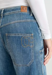 Jean en denim bleu avec un patch en cuir sur la taille, et une poche arrière avec des coutures jaunes et un design unique.