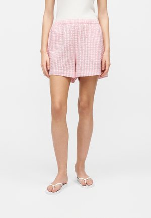 Femeie purtând pantaloni scurți în carouri roz și alb, maiou alb și papuci albi, stând pe un fundal alb simplu.