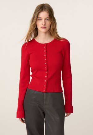 Vrouw met lang blond haar draagt een aangesloten rood gebreid vest met knopen en een zwarte broek, staand tegen een lichte effen achtergrond.