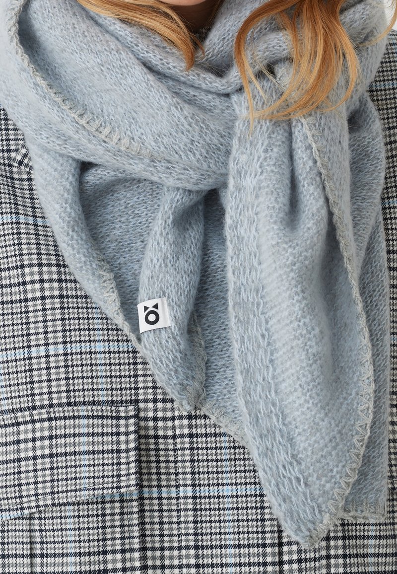 Écharpe en laine gris clair avec étiquette entourée autour du cou, portée sur un manteau à carreaux noir et blanc, vue du torse au menton.