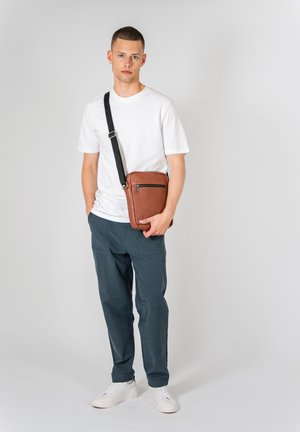Smeđi kožni crossbody ruksak s prednjim zip džepom, podesivim crnim remenom i glatkom teksturom, uparen s bijelom majicom i mornarskim hlačama.