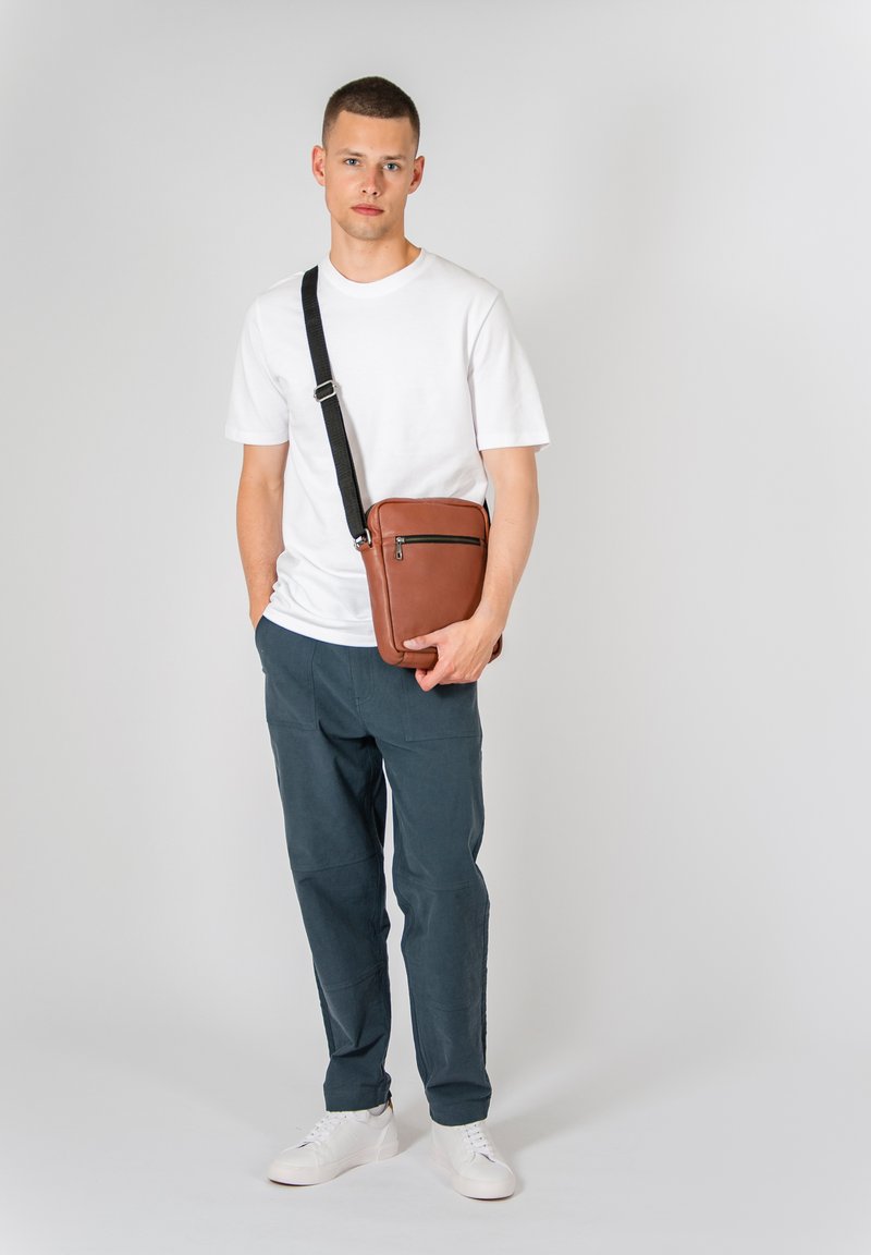 Hnědá kožená crossbody taška s přední zipovou kapsou, nastavitelného černého popruhu a hladkým povrchem, v kombinaci s bílou košilí a námořnickými kalhotami.