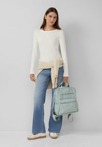 Lichte mintgroene rugzak met twee voorzakken, vastgehouden door een persoon in een wit longsleeve-shirt en lichtblauwe uitlopende jeans.