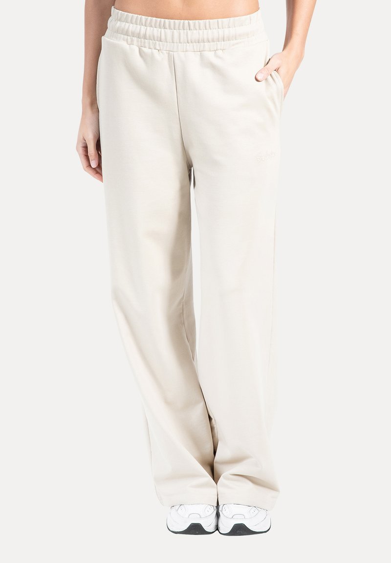 Beige joggingbukser med en glat tekstur, bredt ben-design, elastisk talje og sidelommer, kombineret med hvide løbesko.