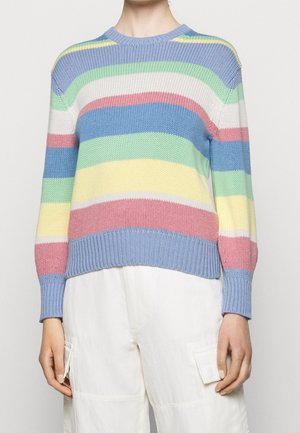 Person trägt einen pastellgestreiften Strickpullover mit blauen, grünen, weißen, rosa und gelben Streifen, kombiniert mit weißen Cargohosen.