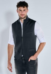 Schwarze, ärmellose Weste mit hohem Kragen, Vorderreißverschluss und zwei Brusttaschen. Getragen über einem weißen Langarmshirt mit glatter Textur.