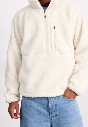 Mand iført hvid fleecepullover med halv lynlås og brystlomme, kombineret med lyseblå jeans, hånd i lommen.