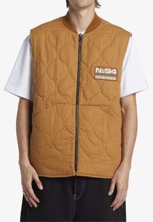 Quilted vest in brown with a zip closure. Features two front pockets and a logo patch. Worn over a white t-shirt with short sleeves.