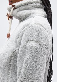 Άτομο που φοράει ανοιχτό γκρι φλις μπουφάν με υψηλό γιακά και κορδόνια με κουμπώματα, με μακριά πλεγμένα μαλλιά ορατά από πίσω.