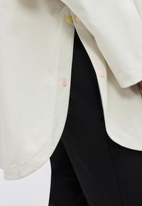 Gros plan sur l'ourlet d'un manteau blanc avec des boutons-pression pastels partiellement ouverts, dévoilant un pantalon noir en dessous.