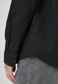 Gros plan sur la main gauche d'une personne, avec le poignet d'une chemise à manches longues noire et texturée, ornée de boutons, et la poche d'un jean en denim gris visible.