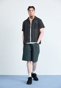 Svart kortermet mønstret skjorte med knapper foran, slitt over en lys grå T-skjorte og kombinert med grønne rutete shorts og svarte joggesko.