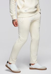 Homme portant un pantalon de jogging écru avec un sweatshirt assorti, associé à des baskets blanches aux semelles marron et aux accents noirs au talon.