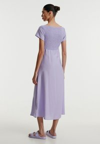 Lila Kleid mit kurzen Ärmeln und gerafftem Rücken. Midi-Länge aus glattem Stoff, mit Seitenschlitzen. Getragen mit passenden lila Sandalen.