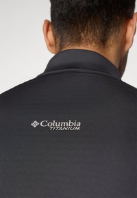 Achteraanzicht van een persoon in een zwarte jas met een opgerichte kraag en het "Columbia Titanium" logo op de bovenrug.