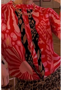 Cárdigan de patrones rojos con gráficos de flores y animales, con botones rosas y un panel negro de acento. Se lleva con pantalones de estampado de leopardo.
