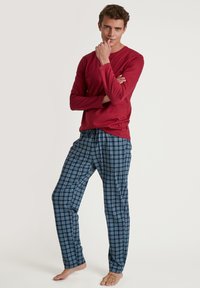 Rode longsleeve shirt gecombineerd met blauwe geruite pyjamabroek met witte en lichtblauwe accenten. Zachte stof, ontspannen pasvorm, op blote voeten.