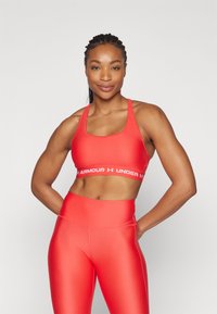 Red sports bra with a wide white band featuring "ARMOUR UNDER" text, paired with matching high-waisted leggings, both in a smooth fabric.