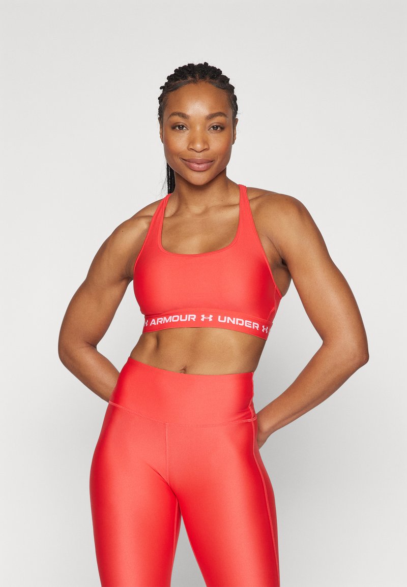 Red sports bra with a wide white band featuring "ARMOUR UNDER" text, paired with matching high-waisted leggings, both in a smooth fabric.
