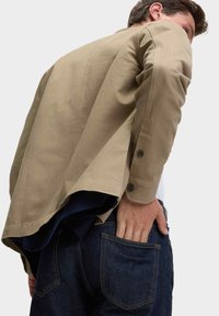 Khaki bunda s knoflíkovým manžetovým rukávem, přetažená přes námořnickou košili, nošená s tmavě modrými džíny. Vyznačuje se elegantní, strukturovanou tkaninou a neformálním designem.