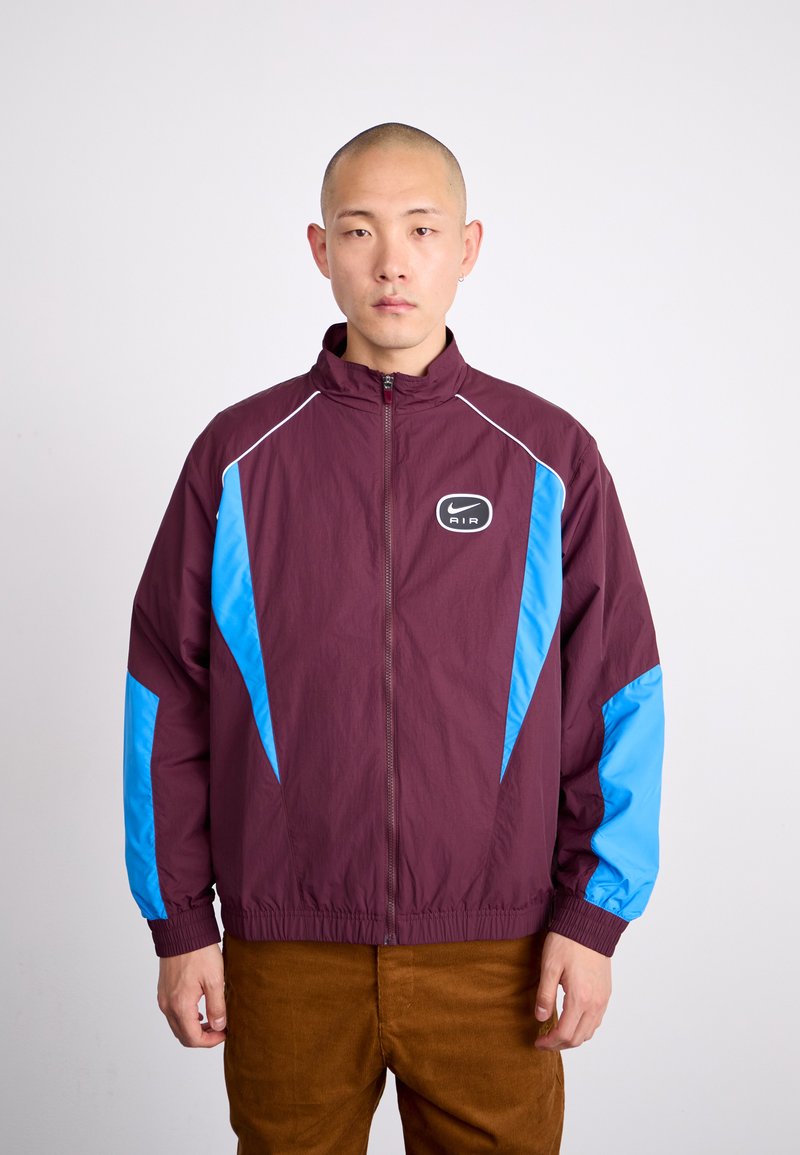 Bordeaux nylonjacka med blå accenter, dragkedjestängning, hög krage och elastiska muddar. Har logotyp på bröstet och diagonala designinslag.