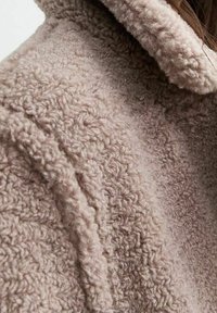 Vezelig beige jasje met een gestructureerde stof en zichtbare naden, dat een warme en gezellige uitstraling heeft.