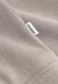 Material bej cu o textură fină, având o etichetă albă cu inscripția "GABBA" cusută în cusătură. Materialul pare moale și flexibil.