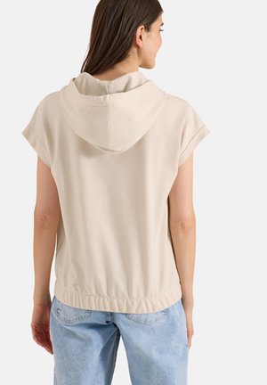 Žena v béžové krátkorukávové mikině s kapucí a světle modrých džínách, zobrazena zezadu na jednobarevném pozadí.