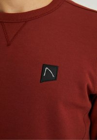 Sweat-shirt bordeaux avec un col ras-du-cou texturé, orné d'un patch carré noir brodé en blanc d'un logo stylisé sur la poitrine.