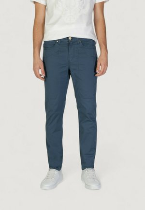 Pantaloni blu a taglio slim con una texture liscia, dotati di tasche frontali e chiusura a bottoni. Indossati con sneaker bianche e una t-shirt bianca con grafica.