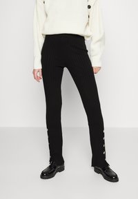 Svarta ribbade byxor med hög midja, med knappdetaljer längs sidan och utsvängd nederdel. Kombineras med en vit stickad tröja och svarta skor.