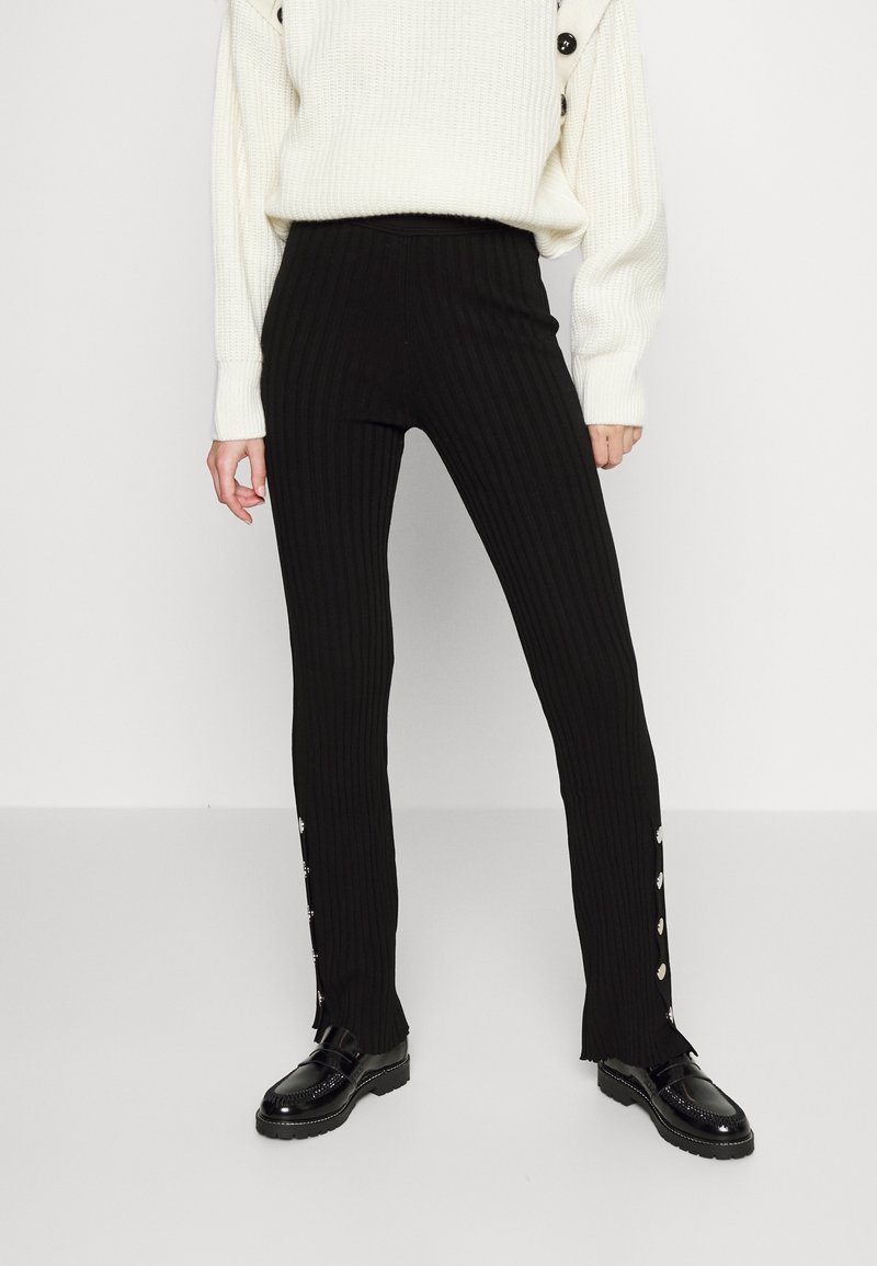 Svarta ribbade byxor med hög midja, med knappdetaljer längs sidan och utsvängd nederdel. Kombineras med en vit stickad tröja och svarta skor.