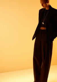 Vrouw in zwarte wijde broek en cropped top met een lange hangende ketting tegen een warme gele achtergrond.