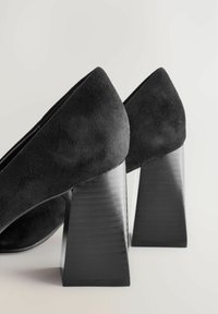 Zapatos de ante negro con tacones en bloque de forma piramidal. La textura es suave, con un acabado mate liso en el tacón.