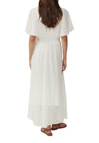 Donna con lunghi capelli castani che indossa un vestito bianco alla caviglia con maniche corte a volant e sandali piatti marroni, vista da dietro.