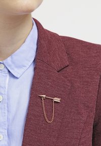 Spilla a forma di freccia in oro rosa con dettaglio a catena, attaccata a un blazer color bordeaux sopra una camicia a colletto azzurro chiaro.