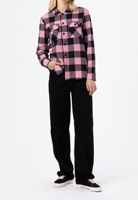 Camicia di flanella a quadri rosa e nera con bottoni frontali, due taschini sul petto e maniche lunghe, abbinata a pantaloni neri e sneakers.