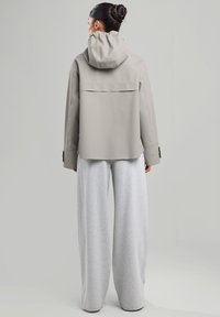Veste imperméable gris clair avec une capuche, présentant une coupe décontractée et des épaules tombantes, assortie à un pantalon de survêtement gris ample.