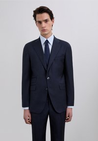 Traje azul oscuro a medida con chaqueta de un solo botón, solapas con muesca, camisa de vestir gris con rayas y corbata azul marino lisa. Tejido suave, ajuste estructurado.