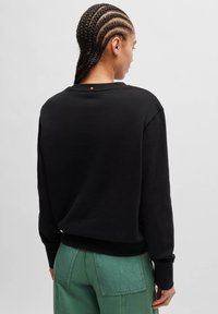 Black sweatshirt with a relaxed fit, ribbed cuffs, and hem. Features a small orange accent at the back neckline. Designed with a smooth texture.