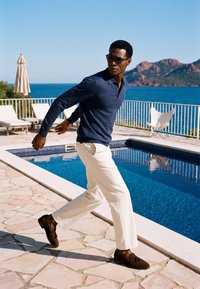 Homme portant des lunettes de soleil, un pull bleu marine, un pantalon blanc et des chaussures marron, marchant près d'une piscine avec l'océan et les montagnes en arrière-plan.