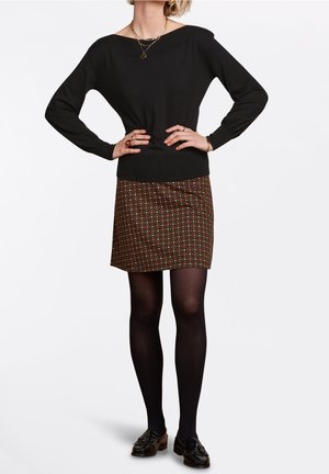 Kobieta w czarnej bluzce z długim rękawem, wzorzystej brązowej mini spódnicy, czarnych rajstopach i czarnych mokasynach z frędzlami, pozująca z rękami na biodrach.