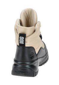 La bota cuenta con una parte superior de nailon beige con un collar acolchado, una parte inferior de goma negra, una superficie texturizada y una lengüeta con marca en la parte trasera.